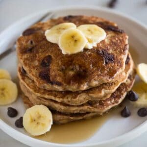 A stack of whole wheat banana bread pancakes on a white plate topped with sliced bananas with chocolate chips on the plate and in the background.