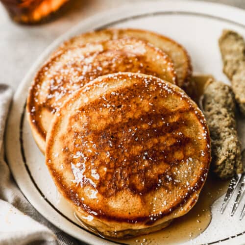 A stack of three homemade whole wheat pancakes topped with powdered sugar and maple syrup on a plate with sausage.