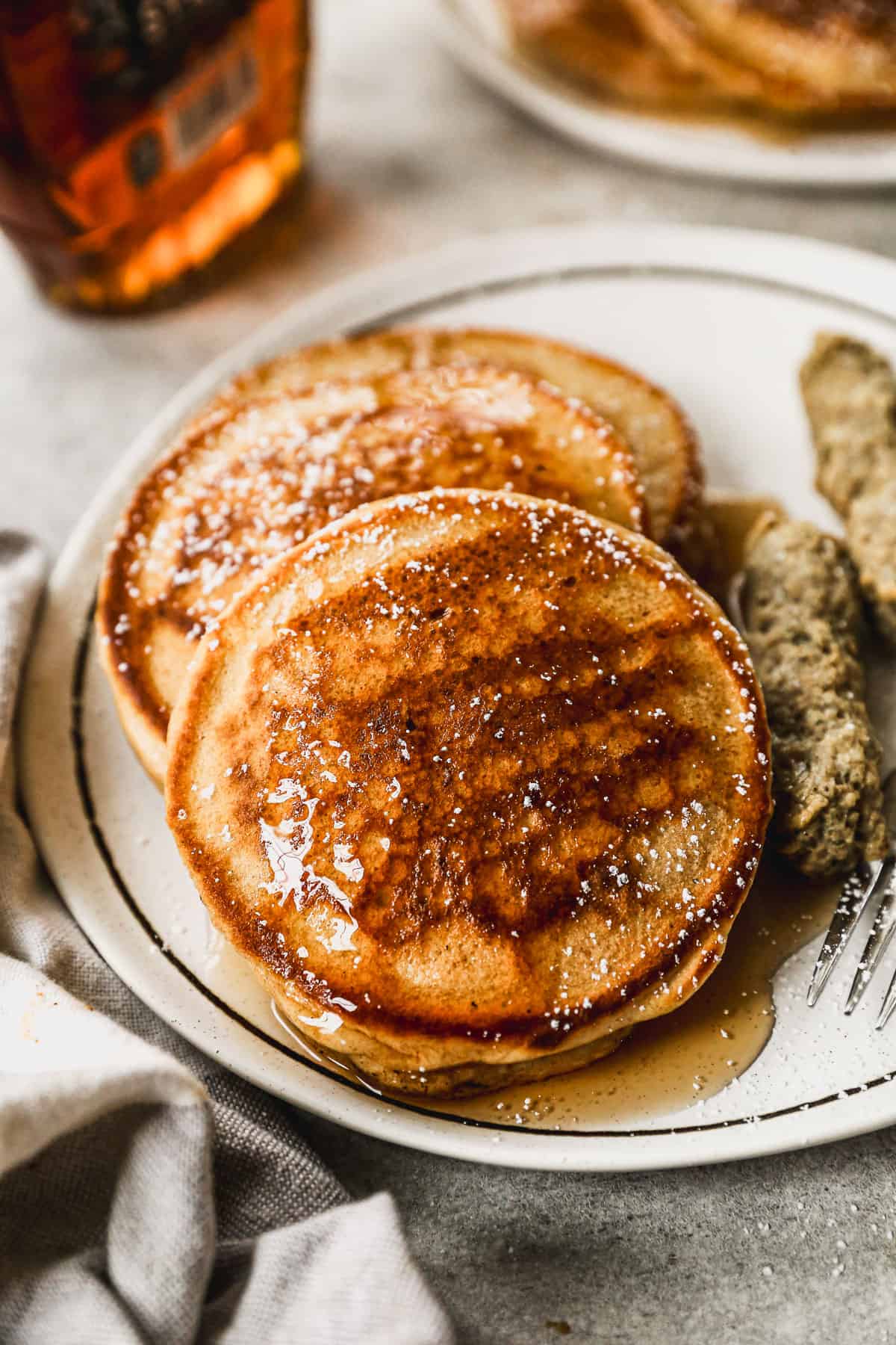 A stack of three homemade whole wheat pancakes topped with powdered sugar and maple syrup on a plate with sausage.